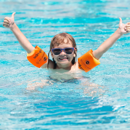 Child in a pool wearing orange arm floats and sunglasses, giving a thumbs-up.